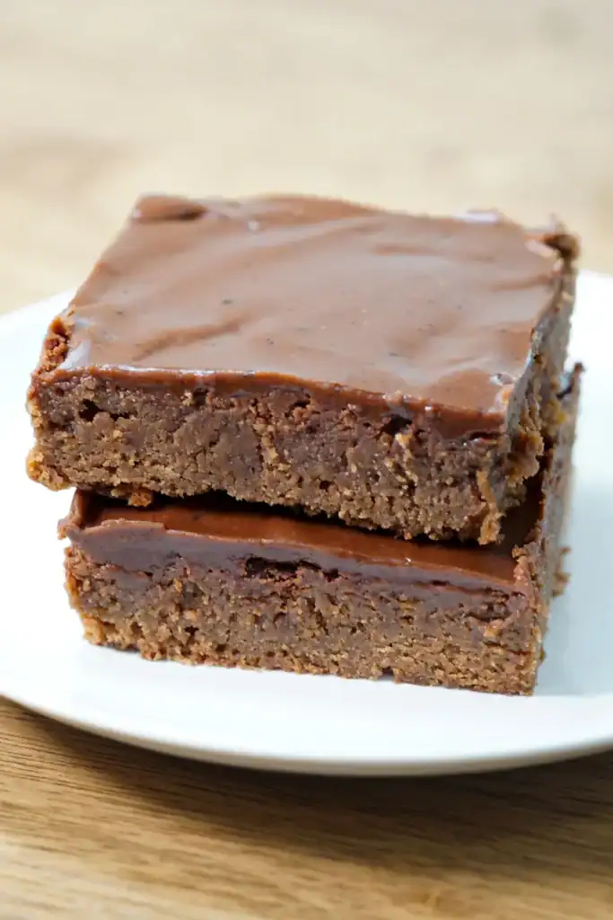 2 squares of iced brownies stacked on top of each other on a white plate.
